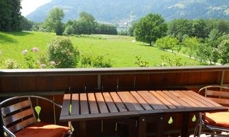 A sunny balcony with a wooden table and two chairs. In the background, green meadows and mountains can be seen.