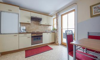 A modern kitchen with light cabinets and a stove. The table is equipped with red chairs, and there are large windows that let in daylight.