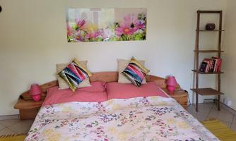 A cozy bedroom with a double bed and colorful cushions. A picture with floral motifs hangs on the wall, and a shelf stands next to the bed.