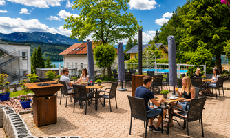 An inviting outdoor area with tables and chairs, where people enjoy drinks. In the background, trees and mountains as well as a blue sky can be seen.