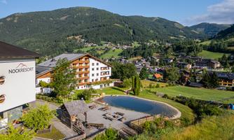 A beautiful view of the Nock resort with a pond and green meadows. In the background, the mountains and the village are visible.