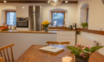 A cozy kitchen with a wooden table and a candle. In the background, modern kitchen appliances and windows with a view outside can be seen.