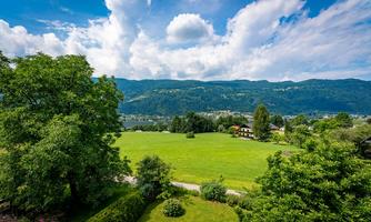 A wide green meadow with trees and some houses in the background. The sky is blue with white clouds and the mountains are visible in the distance.