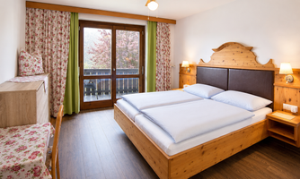 A cozy bedroom with a large double bed; upon request, you can also have 2 single beds. and wooden furniture. The windows offer a view of nature and are framed by bright curtains. with access to the balcony