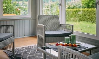 A cozy conservatory with rattan chairs and a table topped with fresh fruit and candles. In the background, a green landscape and mountains can be seen.
