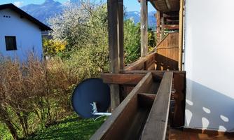 A balcony with wooden elements and a view of the surrounding landscape. In the background, mountains and a clear blue sky can be seen.
