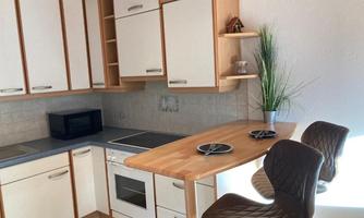 A modern kitchen with light cabinets and a wooden countertop. Two chairs are at a small bar with dishes on it.