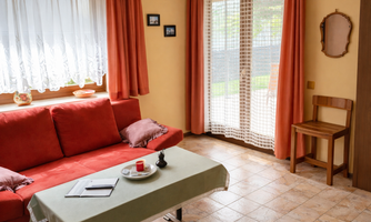 A cozy living room with a red sofa and a small table. Large windows let in plenty of light and provide access to an outdoor area.