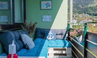 A cozy balcony with a blue seating area and a red table. In the background, green mountains and a clear sky can be seen.