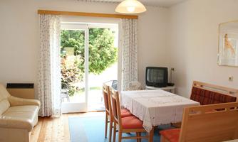 A cozy living space with a dining table, chairs, and a television. The light comes in through the windows, and there is a view of the garden.