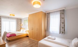 A bright and modern room with two beds, a wooden dresser, and large windows. The decor is simple and inviting, perfect for a pleasant stay.