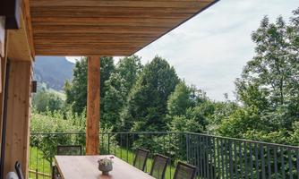 A beautiful balcony with a large wooden table and chairs. In the background, green trees and a clear sky can be seen.