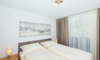 A modern furnished bedroom with a double bed and soft colors. Large windows with curtains let in a lot of light.