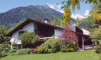 A charming house surrounded by lush greenery and colorful plants. Impressive mountains can be seen in the background.