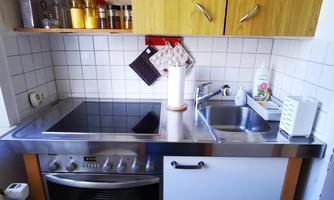 A small, modern kitchenette with a stainless steel surface, stove, and sink. Cooking utensils and spices are hanging on the wall.