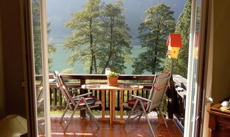 A cozy terrace with two chairs and a table. In the background, there are trees and a beautiful view of a lake.