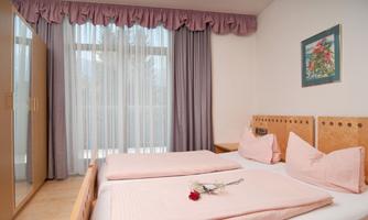 A cozy room with two beds and delicate pink bedspreads. Large windows let in plenty of daylight.