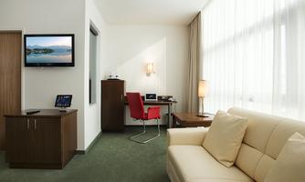 A modern hotel room with a comfortable sofa and a desk. A television is mounted on the wall, and large windows provide plenty of daylight.