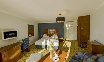 A modern hotel room with a large double bed, a television, and an elegant carpet. There is a cozy seating area with a table and chairs.