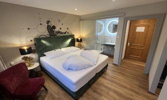 A modern room with a large bed and stylish furniture. The walls are decorated with subtle artwork, and the room offers a cozy seating area.