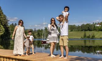 A family stands on a wooden pier by the shore of a calm lake. They are enjoying the sunny day and the beautiful landscape in the background.