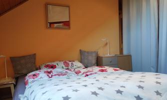 A cozy bedroom with a bed adorned with a colorful duvet and pillows. A mirror hangs on the wall and curtains let gentle light in.