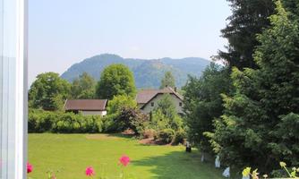 A green meadow with colorful flowers in the foreground and a beautiful view of mountains in the background. There are some houses surrounded by trees.