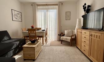 A cozy living room with a gray sofa, a wooden table, and chairs. The large front window lets in plenty of daylight.