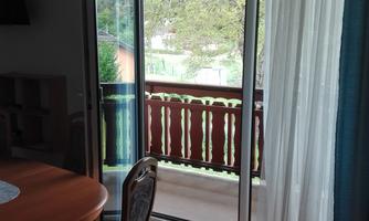 A room with a table and chairs. The open balcony door offers a view of a balcony and a green outdoor area.