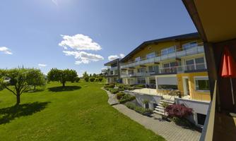 A modern building with a balcony and large windows, surrounded by green grass and trees. The sky is blue with some white clouds.