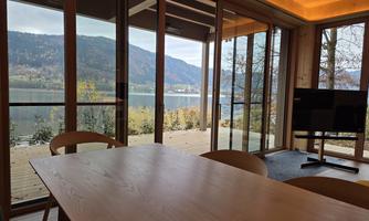 A modern room with a large table and a view of a clear lake. The windows overlook the surrounding mountains and nature.