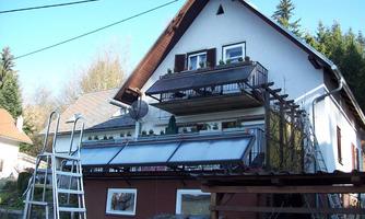 A house with a balcony and a ladder stands in the foreground. In the background, trees and a clear sky can be seen.