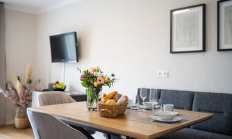 A modernly furnished dining area with a wooden table and atmospheric floral decoration. In the background, there is a television and a cozy couch.
