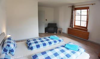 A bright room with two single beds in blue check patterns. There is a window overlooking the outside and a cozy armchair in the room.