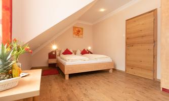 A cozy bedroom under the roof with a double bed and red cushions. The walls are painted in a soft color, and there is a wooden chair as well as a door leading to the hallway.