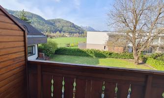 A balcony with a view of green meadows and mountains in the background. The sky is clear and the sun is shining.