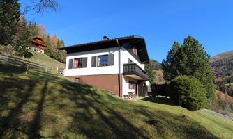 A cozy house on a green hill, surrounded by trees. The sky is clear and blue, which highlights the peaceful landscape.