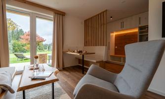 A modern, bright room with a comfortable armchair and a table. Large windows offer a view of the green landscape outside.