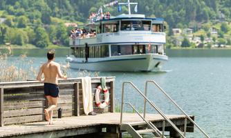 A man walks along the shore of a lake as a pleasure boat with passengers passes by. Surrounded by green hills, the scene offers a relaxed summer atmosphere.