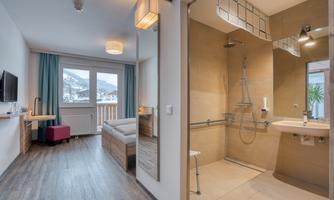 A modern hotel room with a comfortable bed and an adjoining bathroom. Large windows provide a view of the surroundings and plenty of daylight.