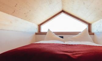 A cozy bedroom with a bed and two pillows. The windows offer a view of the sky and provide plenty of natural light.