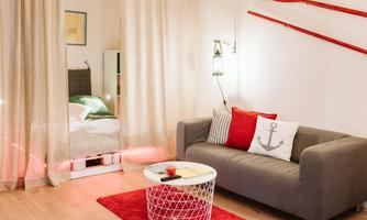 A cozy living room with a gray sofa and colorful cushions. In the background is a bed and some decorative elements.