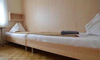 A simple room with two beds and a small table. The walls are light and the floor is made of wood.