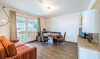 A bright and modern living kitchen with a cozy sofa and a dining table. Large windows provide plenty of daylight.