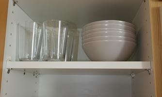 A kitchen cabinet with various glasses and dishes. Above are tall glasses, and beneath are bowls and plates arranged in soft colors.