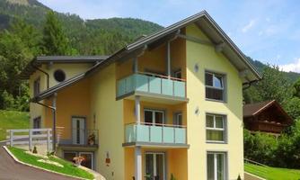 A modern, yellow residential building with balconies and large windows. It is situated in a green, mountainous area.