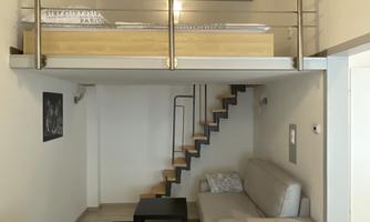 A modern living room with a sleeping loft and a staircase. The room is designed to be bright and friendly.