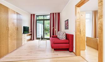 A modern living room with a red sofa and a television. Large windows provide a lot of daylight and a view of the outdoors.