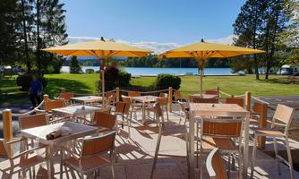 An inviting outdoor area with tables and sun umbrellas. In the background, a calm lake and a green meadow can be seen.