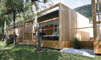 Modern wooden cabins in a green setting with mountain views. The terraces are invitingly furnished with comfortable seating.